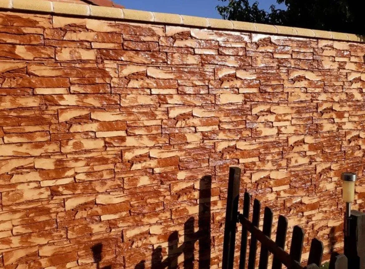 Muro de ladrillo con acabado texturizado marrón rojizo bajo un cielo azul, parcialmente oculto por una valla.