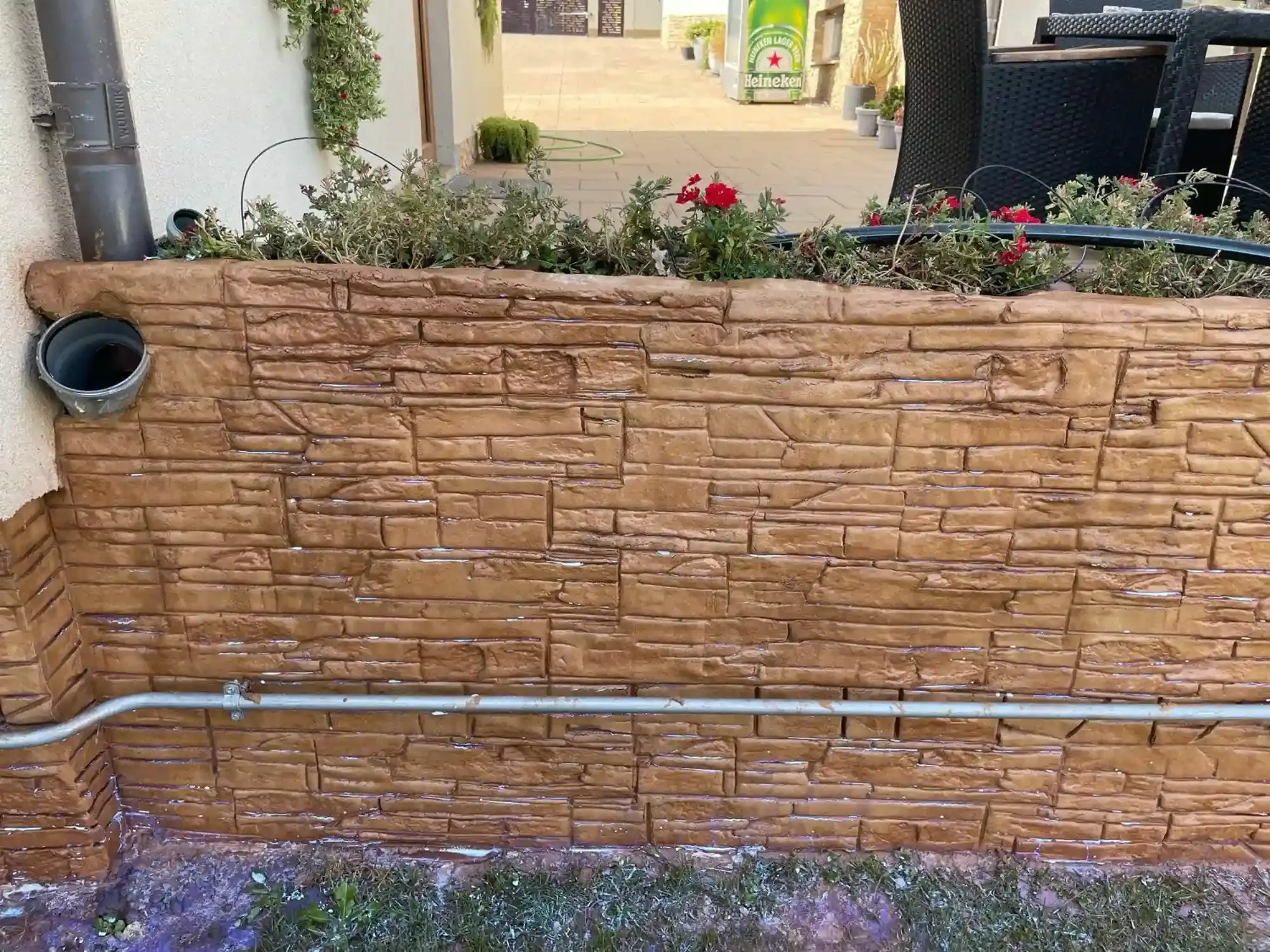 Pared de ladrillo marrón con plantas en la parte superior, un canalón y una tubería metálica que recorre la base de la pared.