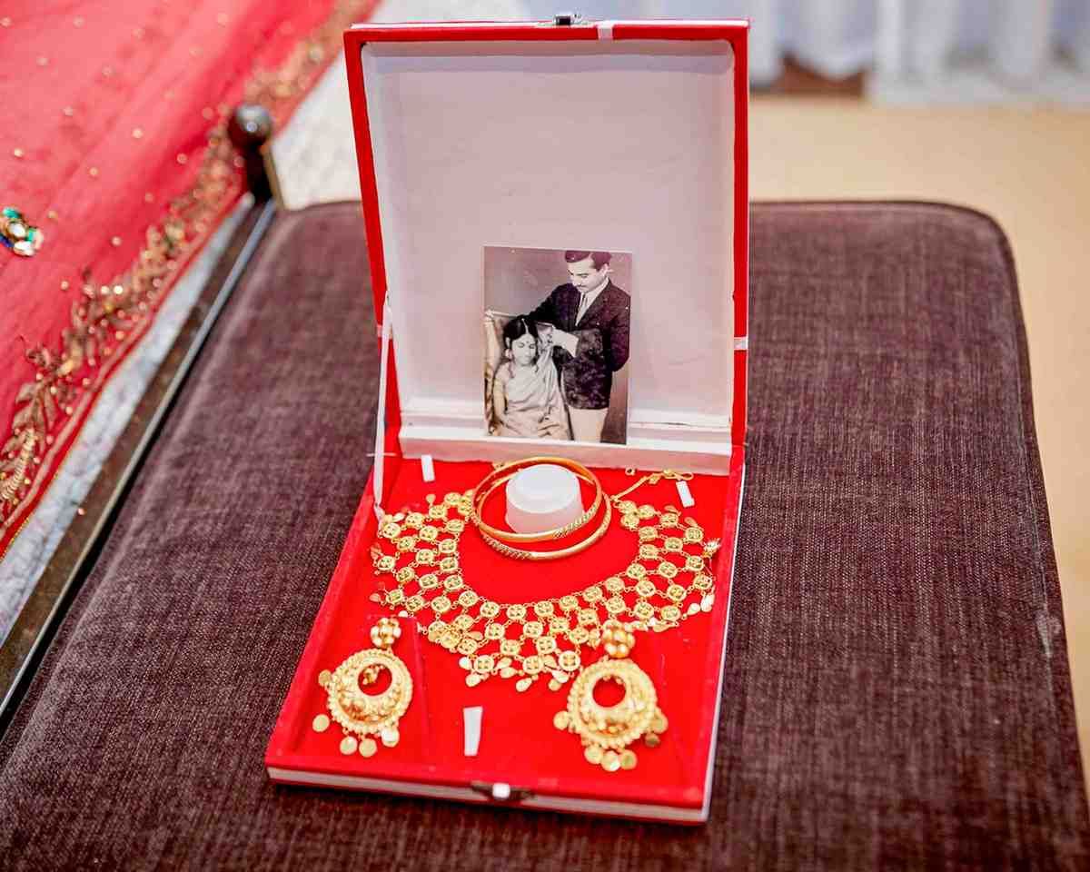 Caja de joyería roja con collar y pendientes de oro, abierta sobre una superficie marrón con una pequeña foto en la tapa.