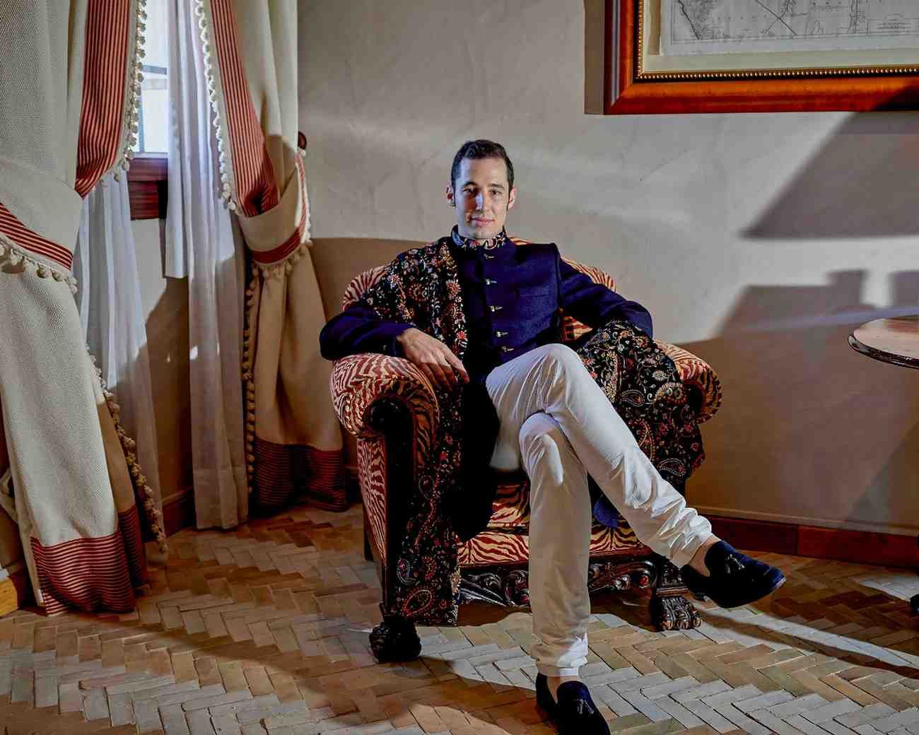 Un hombre sentado en un sillón estampado junto a unas cortinas, vestido con una chaqueta oscura y pantalones.
