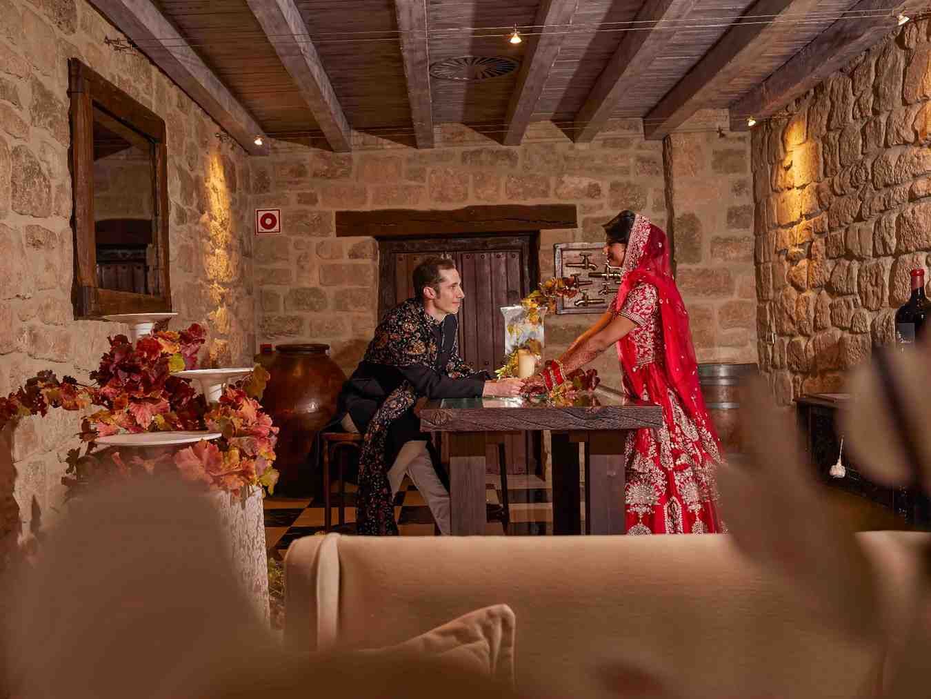 Dos personas en una habitación rústica de piedra, una vestida de negro y la otra de rojo, extendiendo los brazos.