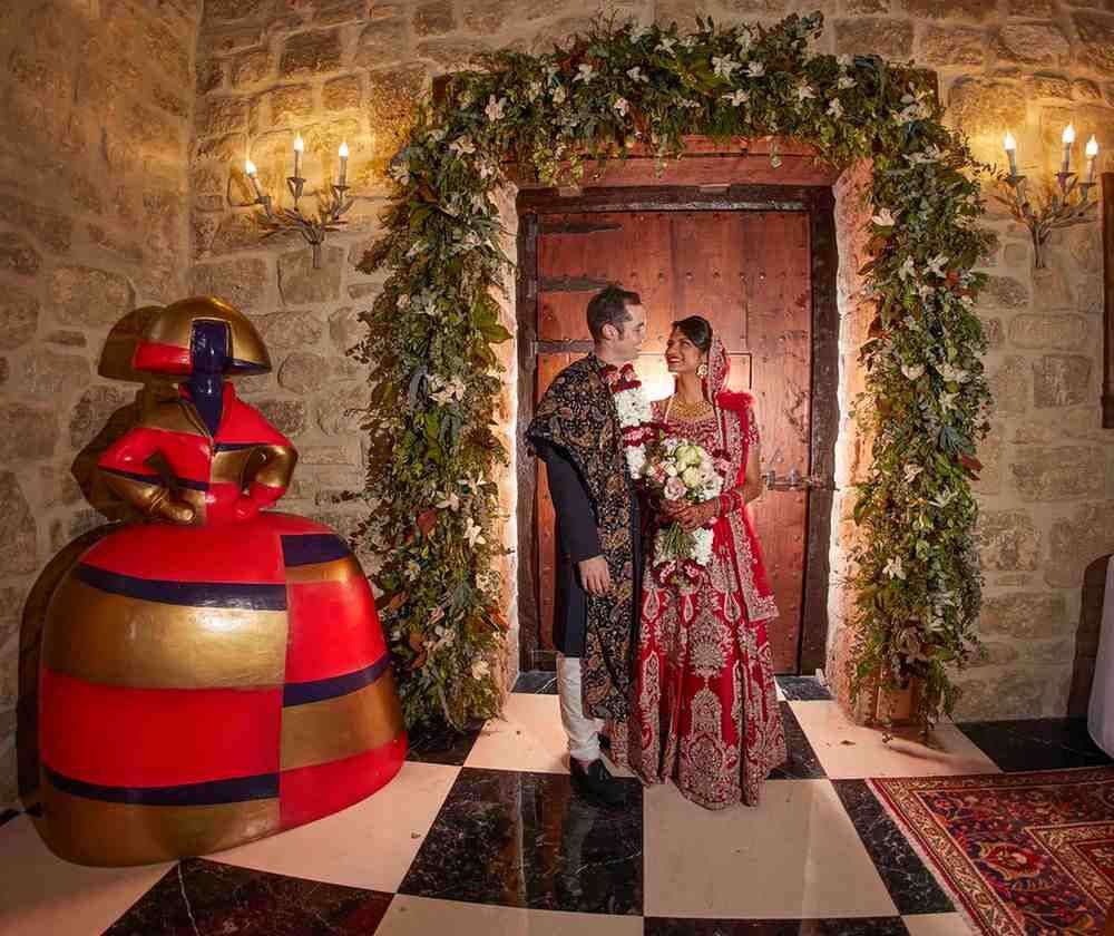 Una pareja vestida de gala posa en el umbral de una puerta de piedra, enmarcada con vegetación y velas.