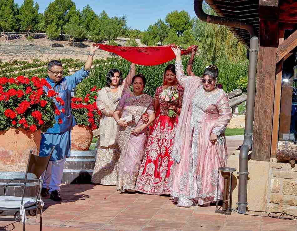Grupo de personas con atuendo formal posando al aire libre bajo un toldo rojo, sosteniendo ramos de flores y sonriendo.