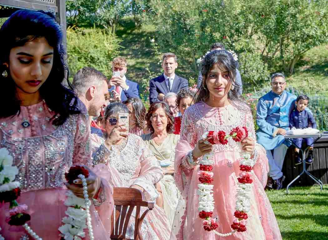 Niños con vestidos rosas caminando al aire libre en una ceremonia en el jardín, con invitados sentados detrás de ellos.