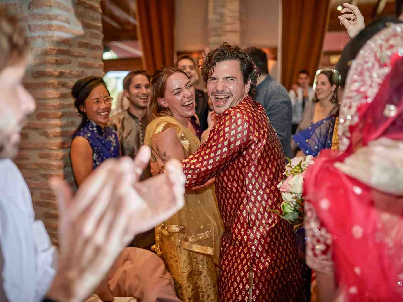 Invitados bailando y aplaudiendo en una festiva celebración en interiores, con una pareja sonriente vestida de dorado.