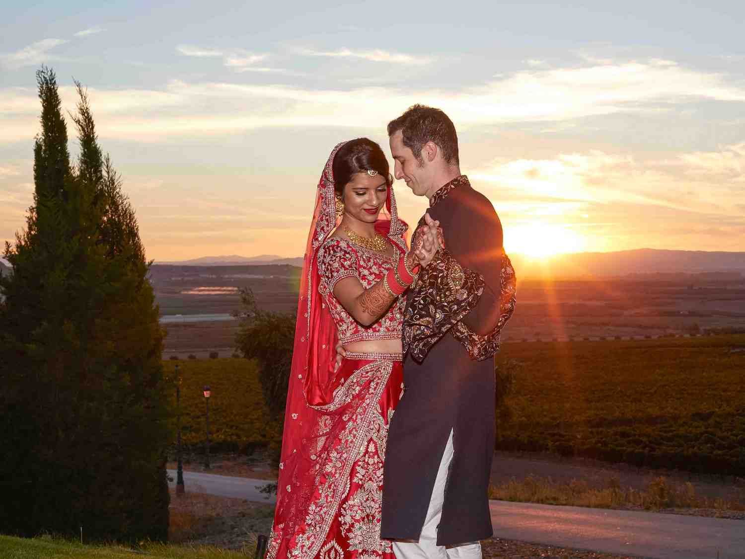 Una pareja vestida con atuendos tradicionales rojos y oscuros se abraza al atardecer en un pintoresco entorno al aire libre.