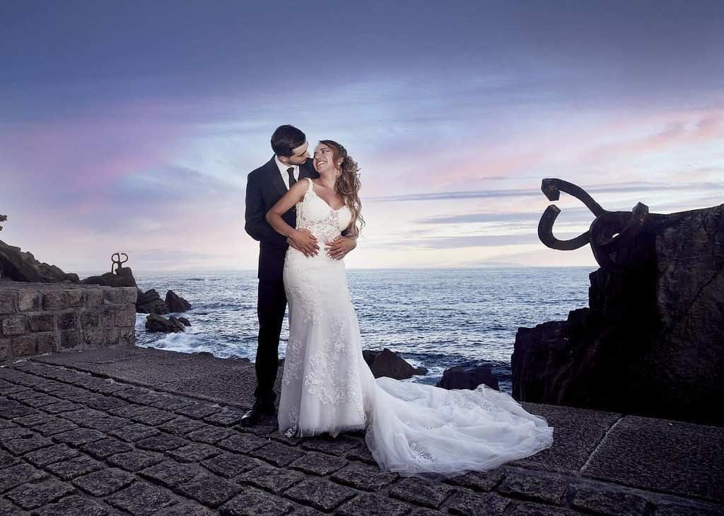 Los novios se abrazan en una terraza junto al mar al atardecer, con las olas del océano y un cielo rosa azulado de fondo.