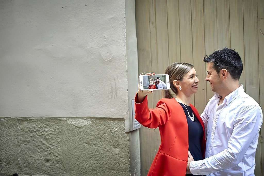 Dos personas sonriendo y posando para una selfie junto a una pared, una con un blazer rojo y la otra con una camisa blanca.