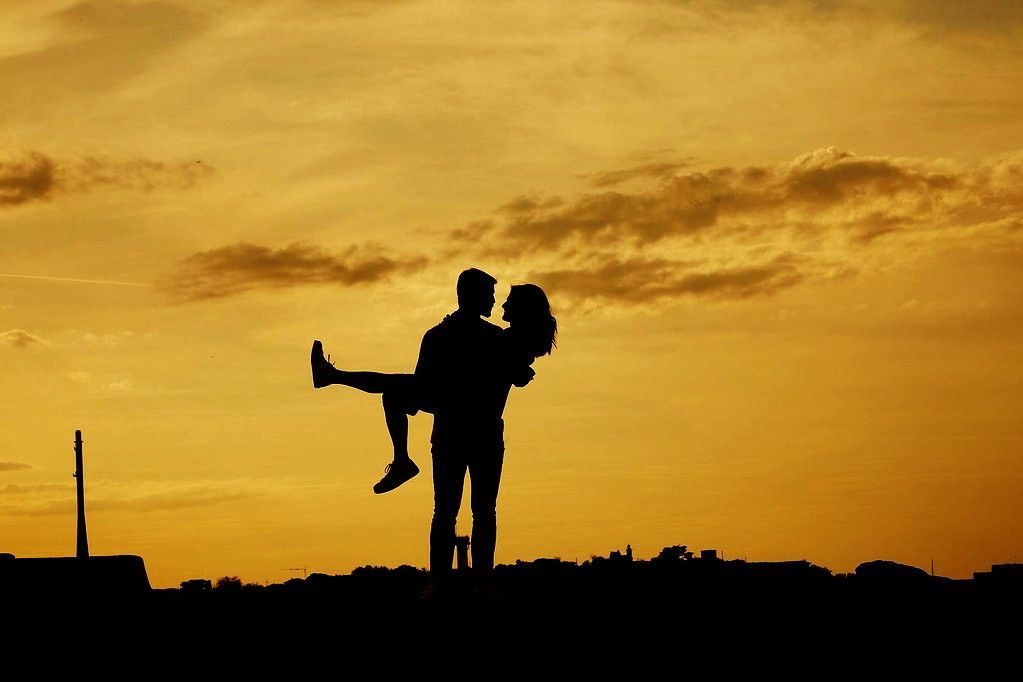 Silueta de dos personas al atardecer, una alzando a la otra contra un cielo dorado.