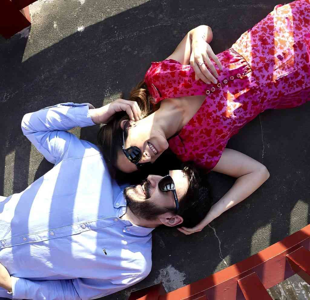 Dos personas tumbadas en la acera, con gafas de sol y ropa de estampados llamativos, con un banco rojo cerca.