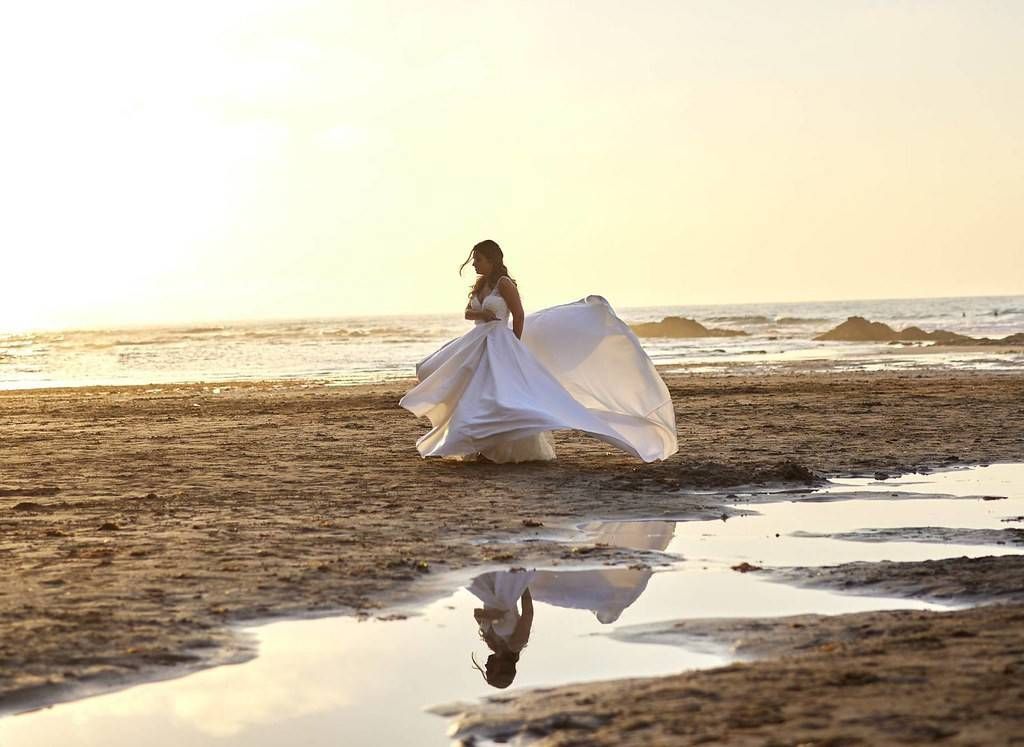 Una persona con un vestido blanco vaporoso camina por la playa al atardecer, reflejada en una poza de marea.