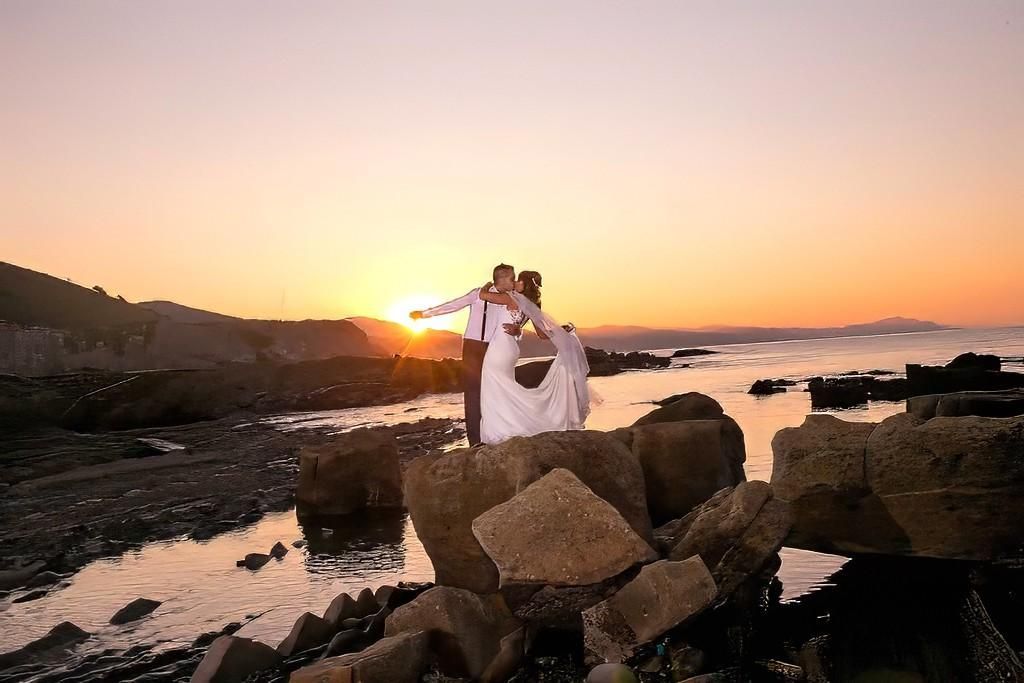 Pareja vestida de blanco de pie sobre rocas al atardecer junto al agua, con cielo anaranjado y niebla.