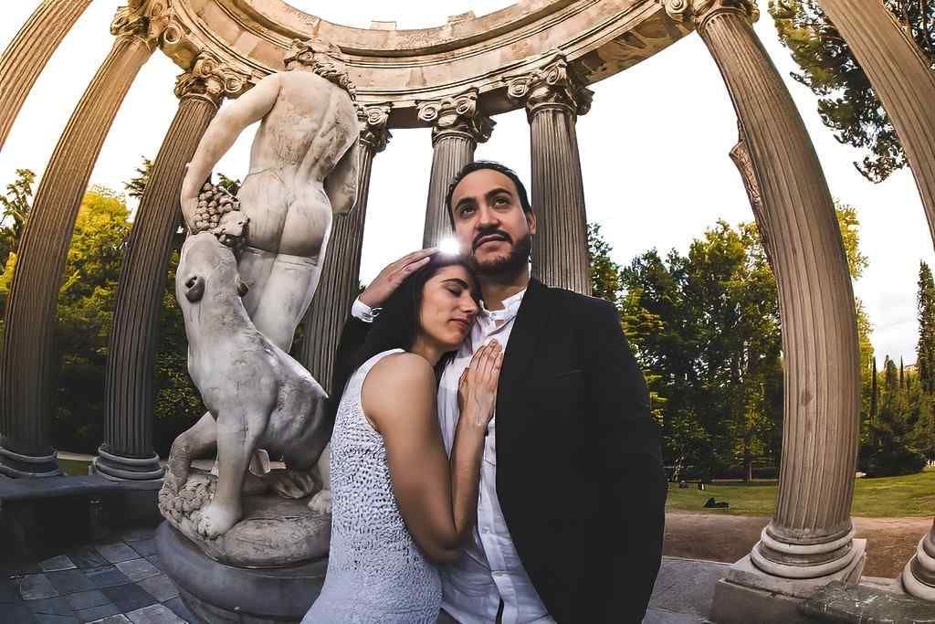 Pareja posando bajo un arco de piedra ornamentado con una estatua en un patio ajardinado.