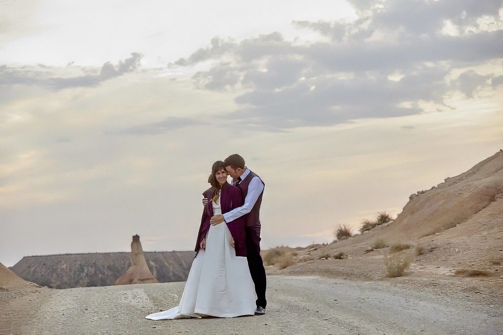 Una pareja se abraza en un paisaje rocoso al atardecer, con acantilados y un cielo nublado de fondo.