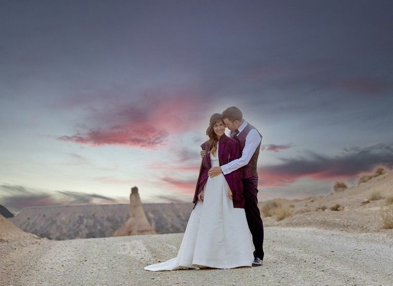 Pareja abrazada vestida de blanco y burdeos en un paisaje desértico al atardecer.
