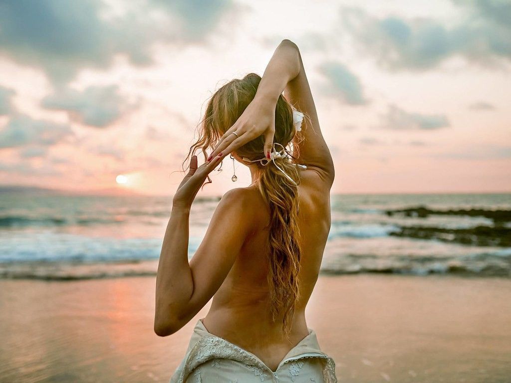 Una persona con una blusa blanca en una playa al atardecer, estirándose con los brazos en alto contra el océano.