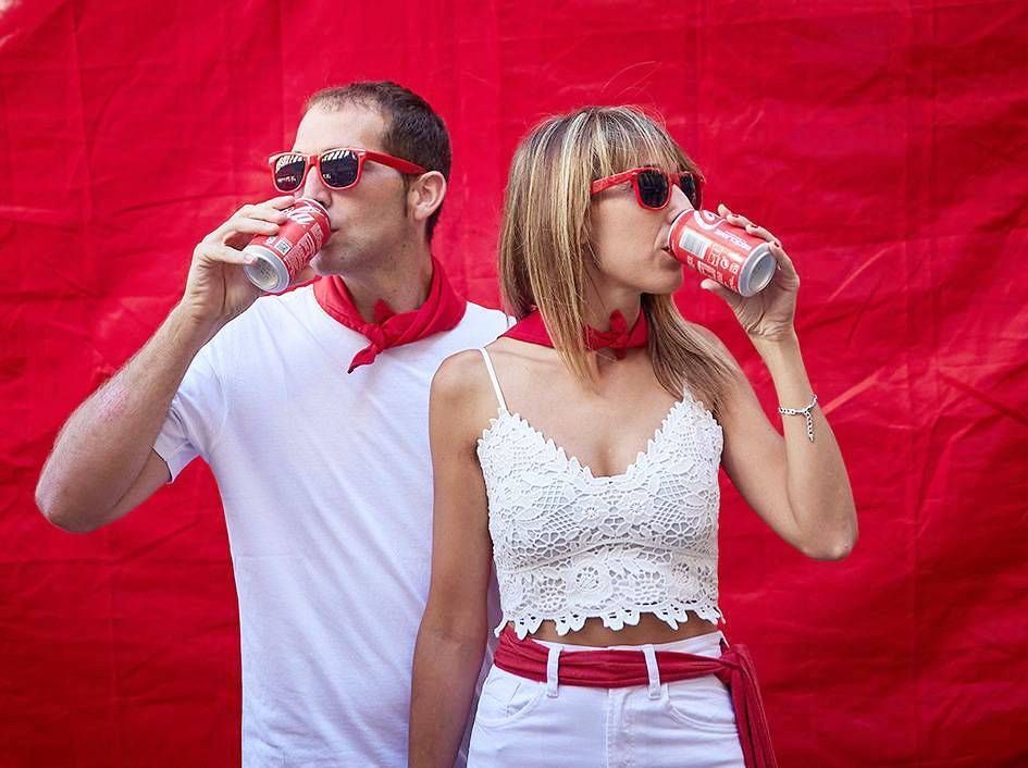 Dos personas vestidas de blanco beben de latas con un fondo rojo brillante.