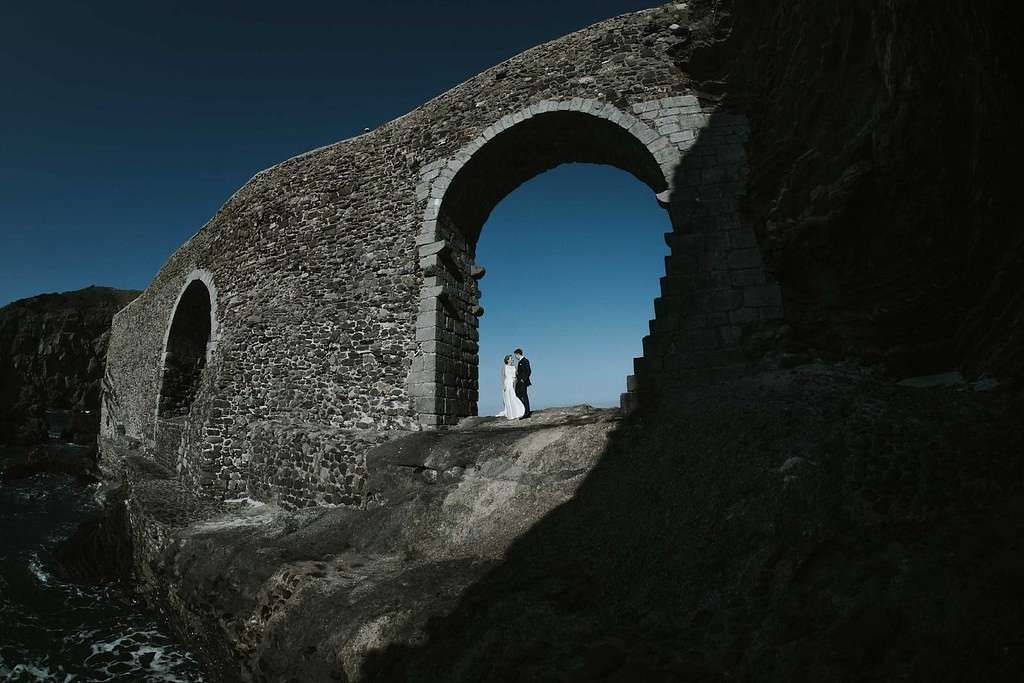 Ruinas de un arco de piedra contra un cielo azul, con dos personas de pie en un sendero iluminado por el sol.