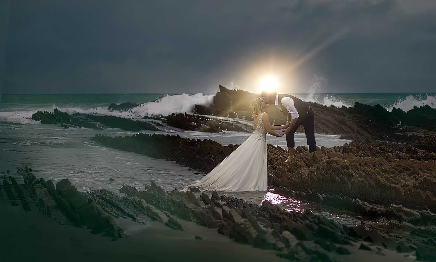 Novios en la orilla rocosa al atardecer, con el viento ondeando el largo vestido blanco de la novia.