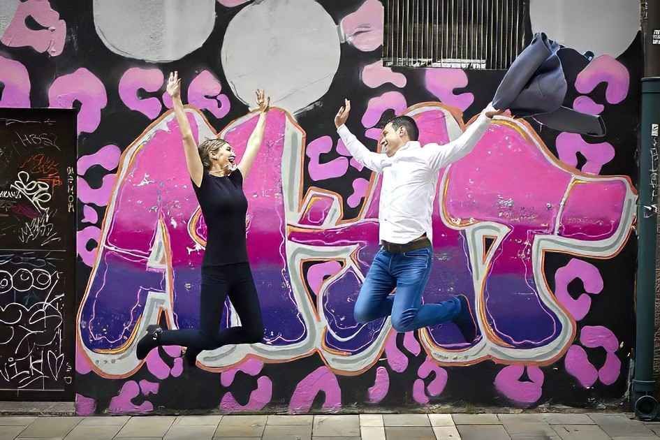 Dos personas saltando frente a un grafiti rosa y negro en una pared.