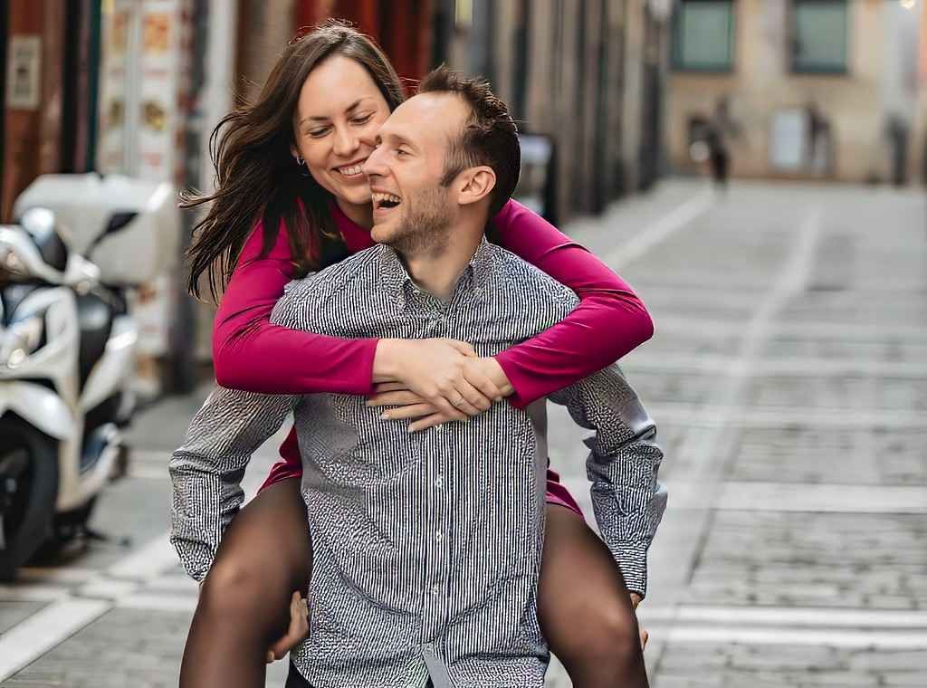 Una pareja sonríe mientras se llevan a cuestas en una calle de la ciudad, con un patinete al fondo.