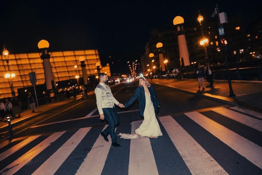 Novios tomados de la mano en un sendero del jardín, vestidos con trajes de boda color marfil y sonriendo.