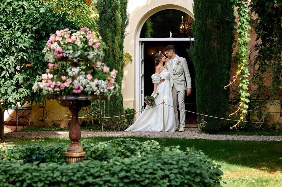 Los novios posan en un patio ajardinado con arreglos florales rosas y arcos cubiertos de hiedra.