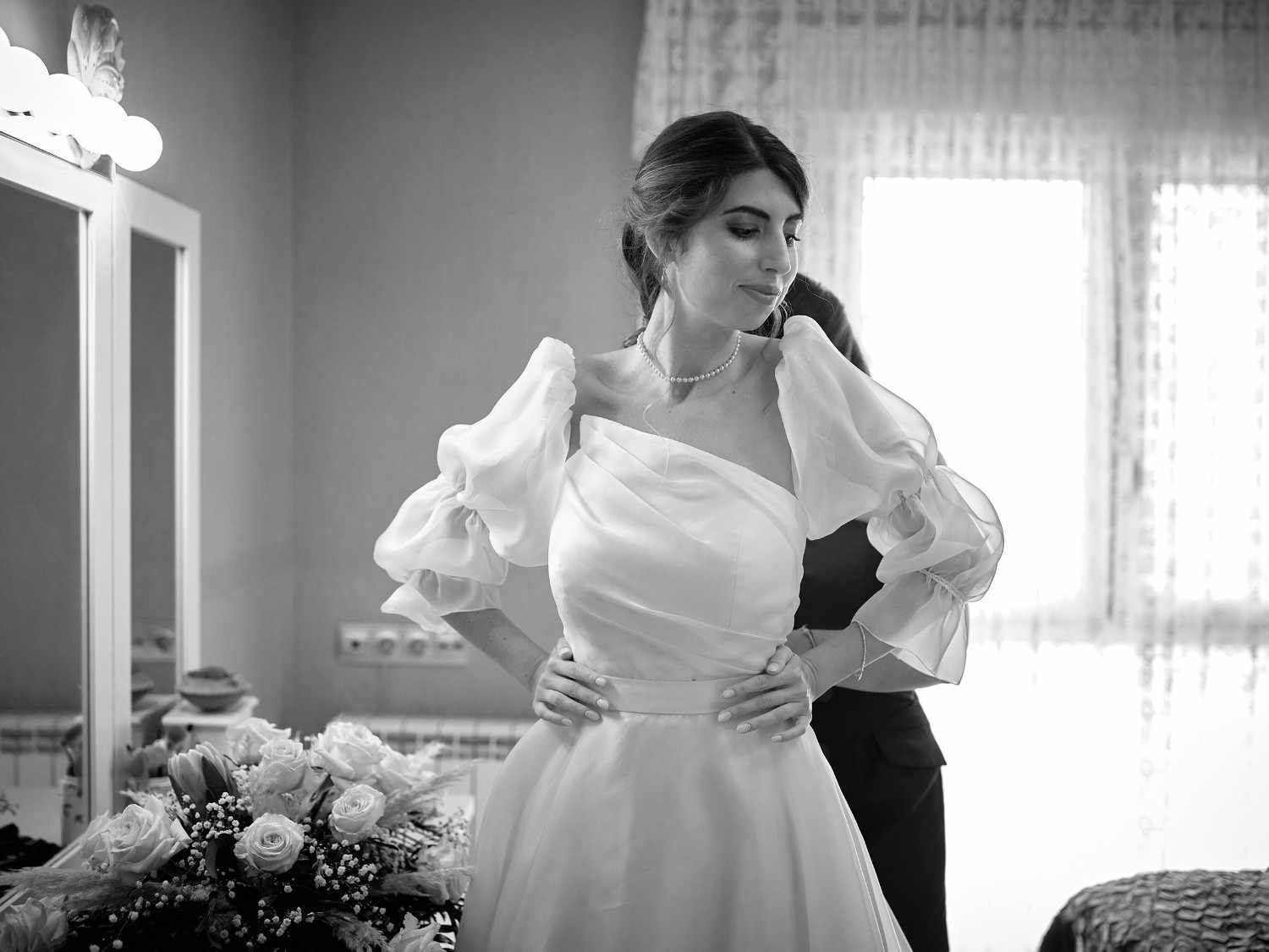 Novia con vestido blanco ajustándose el vestido en una habitación luminosa, con una persona detrás de ella y flores cerca.