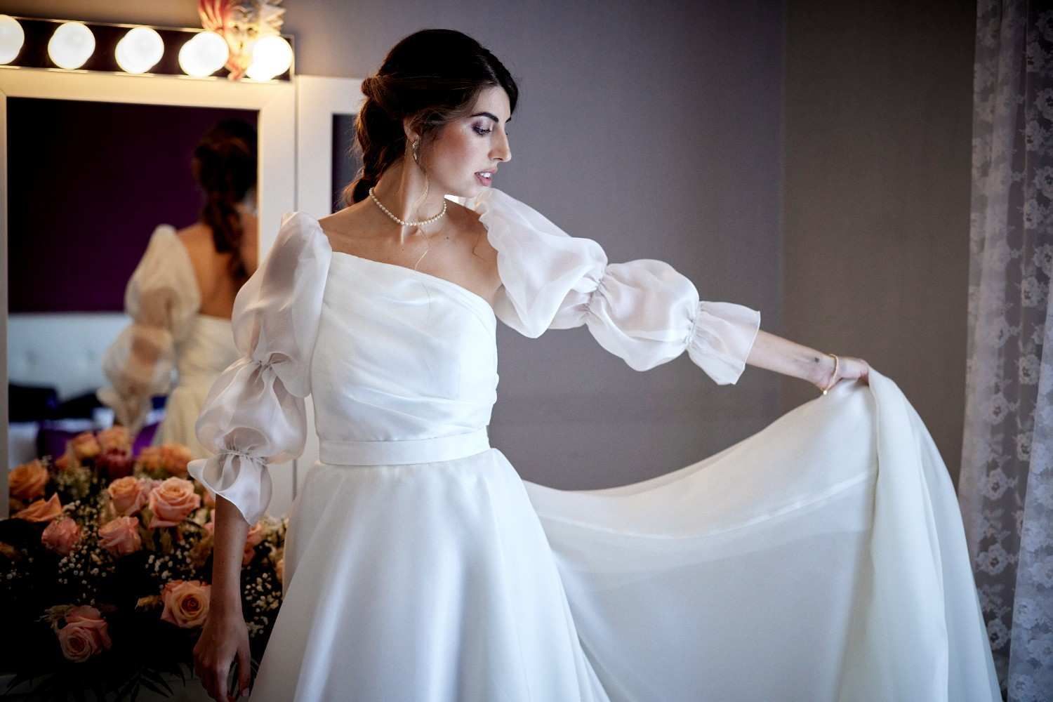 Novia con vestido blanco sosteniendo una cola ondeante en un vestidor.