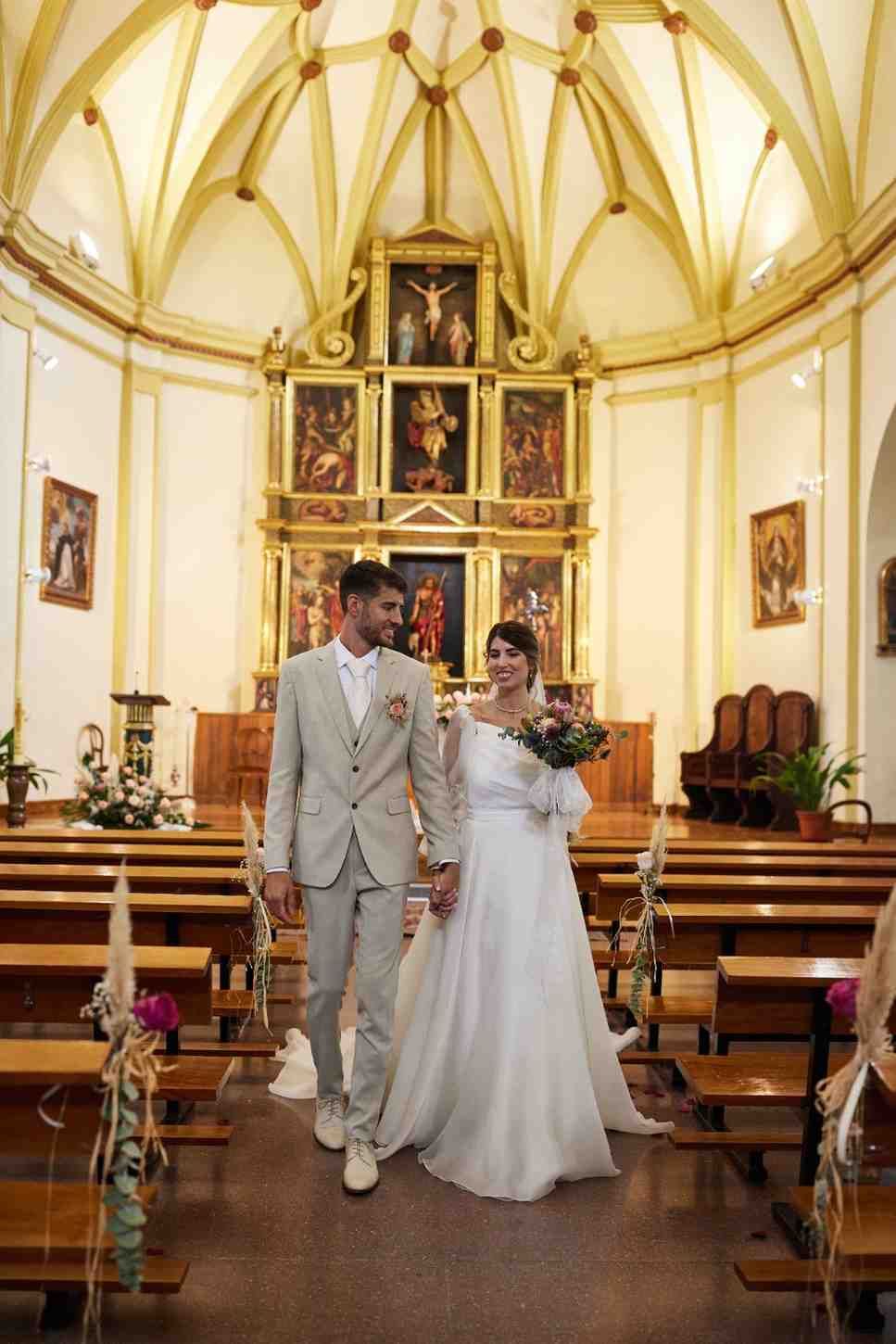 Novios de pie en el pasillo de una iglesia frente a un altar dorado.