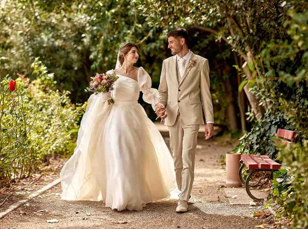 Novios tomados de la mano en un sendero del jardín, vestidos con trajes de boda color marfil y sonriendo.