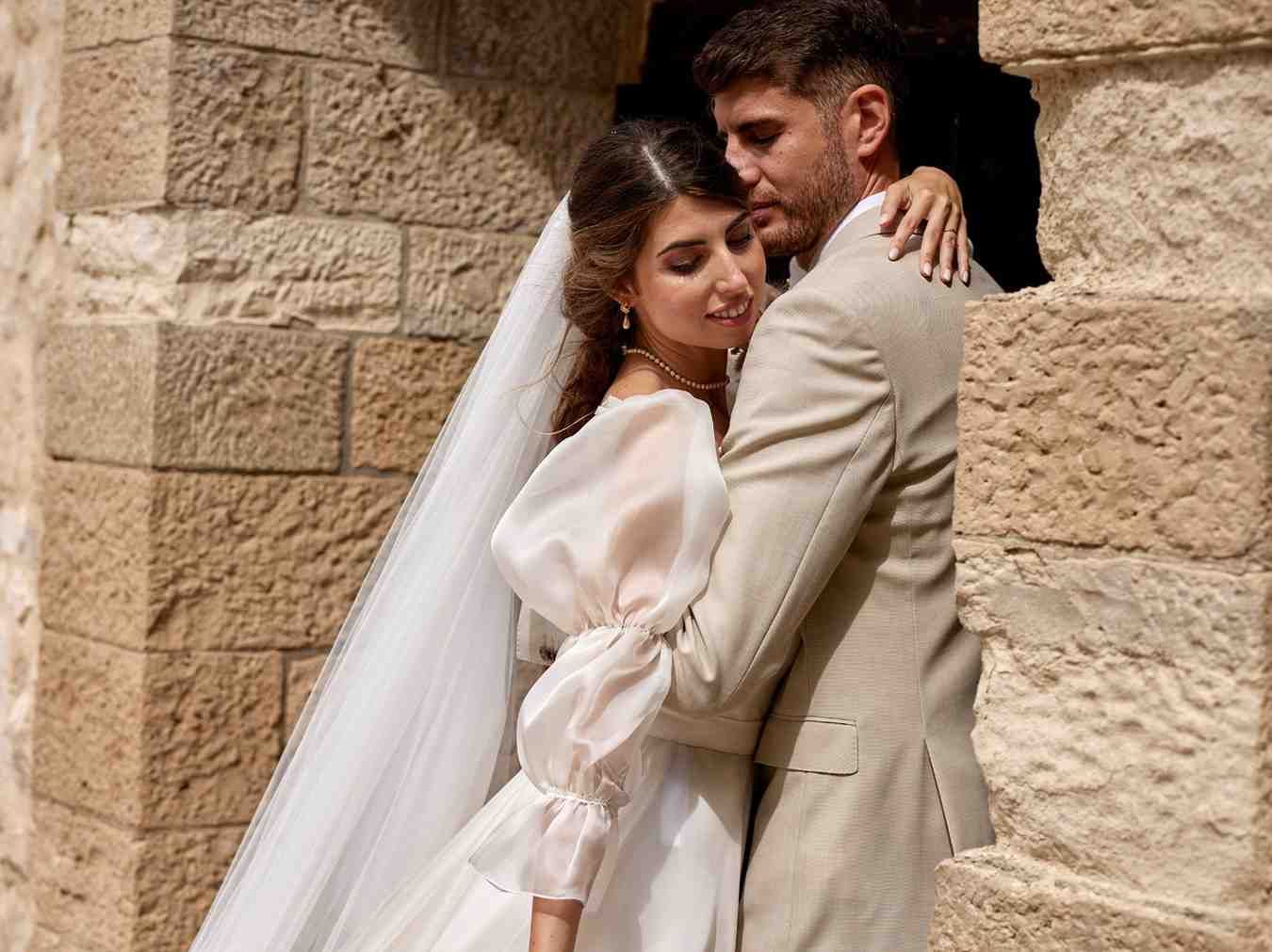 Novios abrazándose con sus trajes de boda contra un muro de piedra.