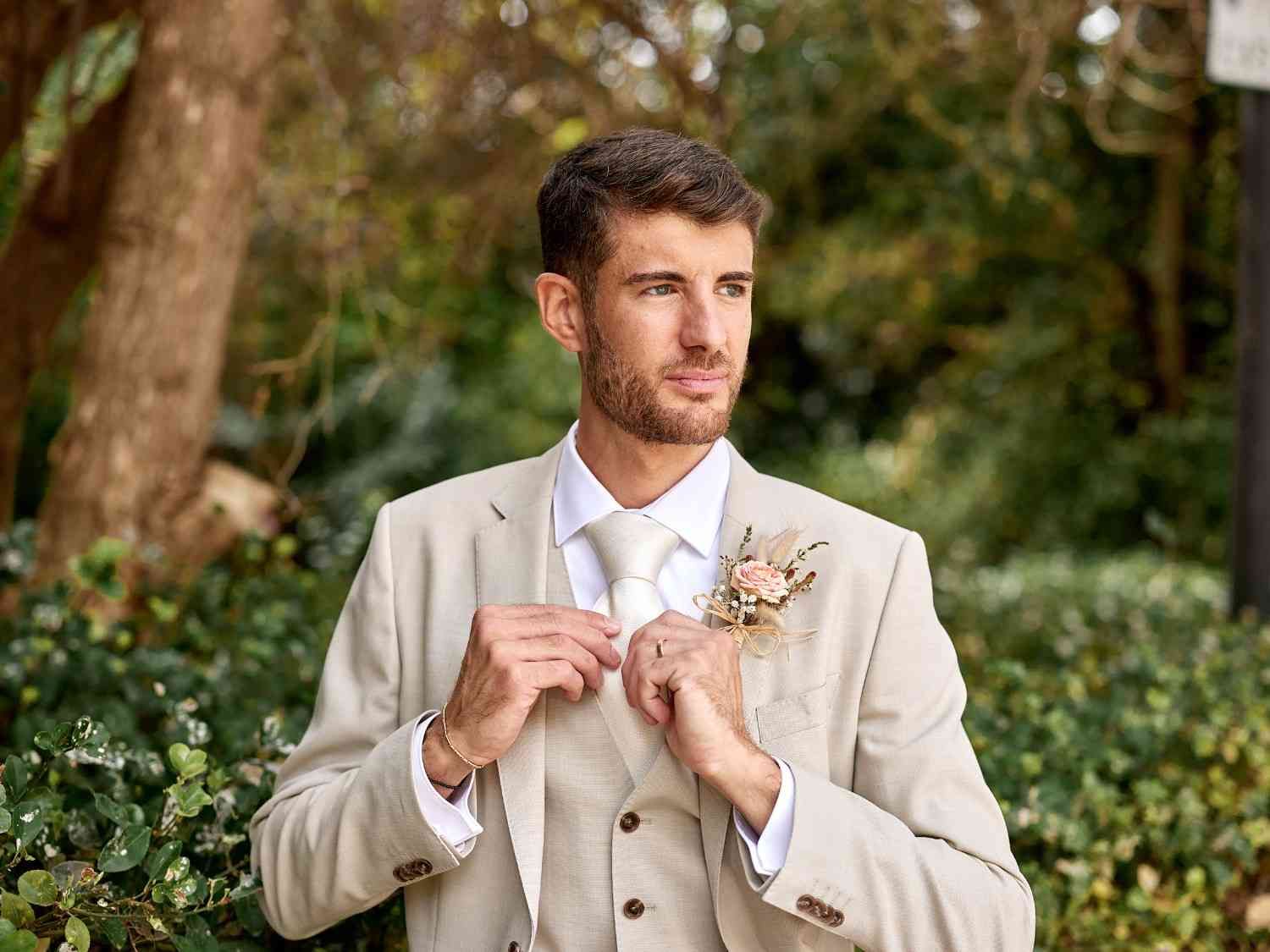 Un hombre con un traje beige se ajusta la corbata al aire libre, con vegetación y árboles al fondo.
