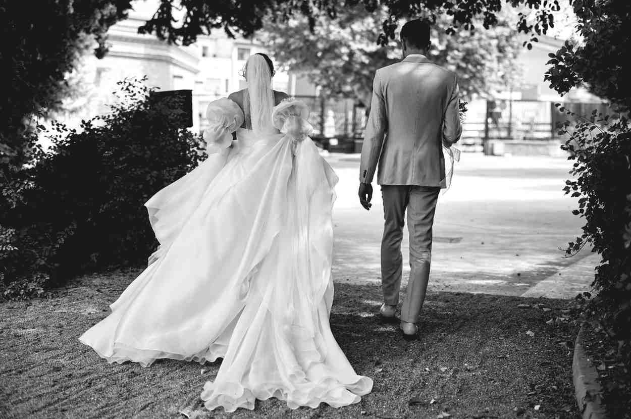 Una novia con un largo vestido blanco y un hombre con traje se alejan caminando por un sendero bordeado de árboles.