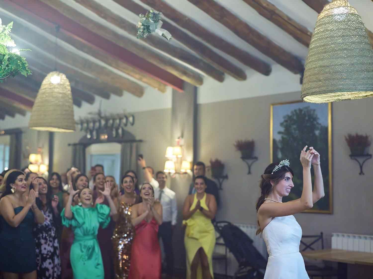La novia, vestida de blanco, lanza el ramo a los invitados que la vitorean en una recepción nupcial en el interior.