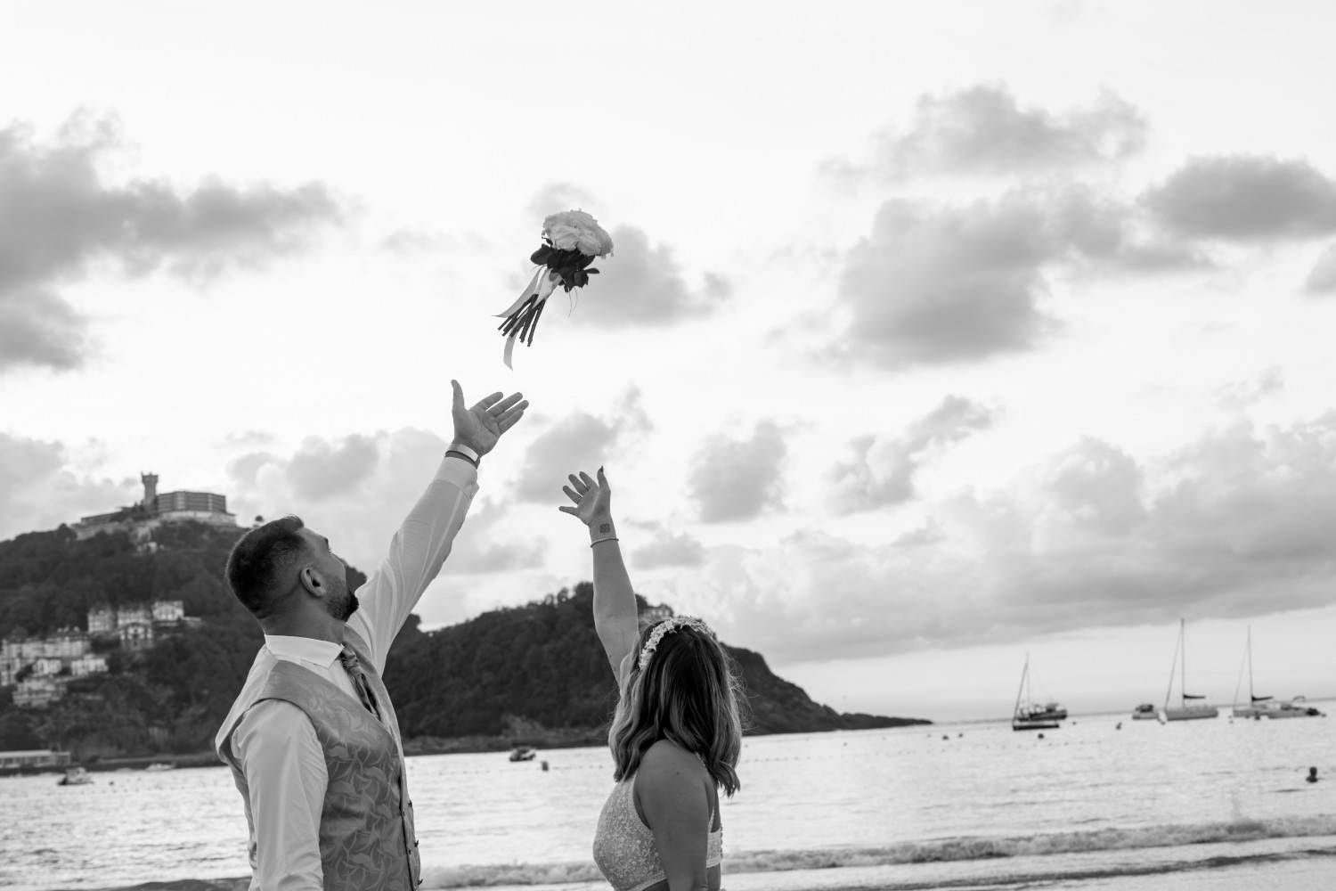 Dos personas lanzan un ramo de flores al aire en una playa con barcos y un cielo nublado de fondo.