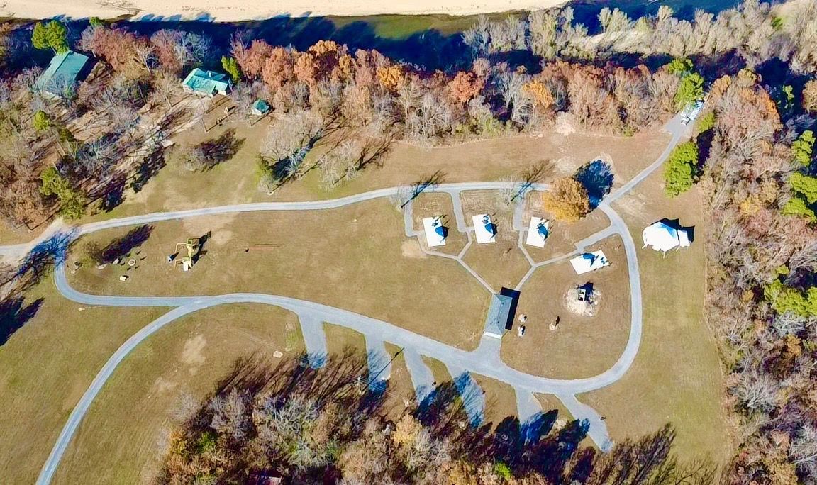 Aerial view of a clearing with small white buildings, paths, and surrounding trees. A river is in the background.
