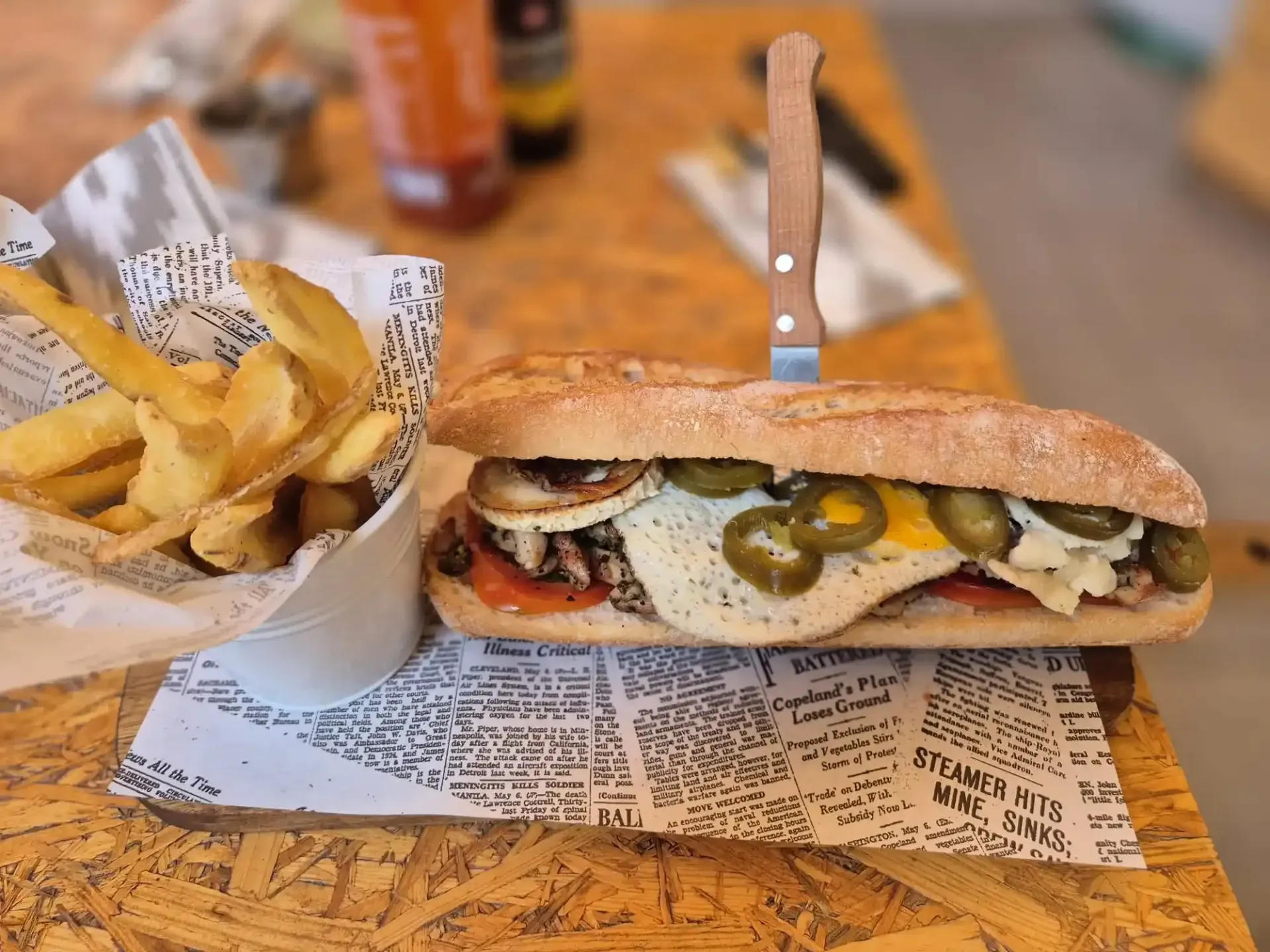 Sándwich con aderezos y patatas fritas sobre una mesa de madera, cuchillo insertado en el sándwich.