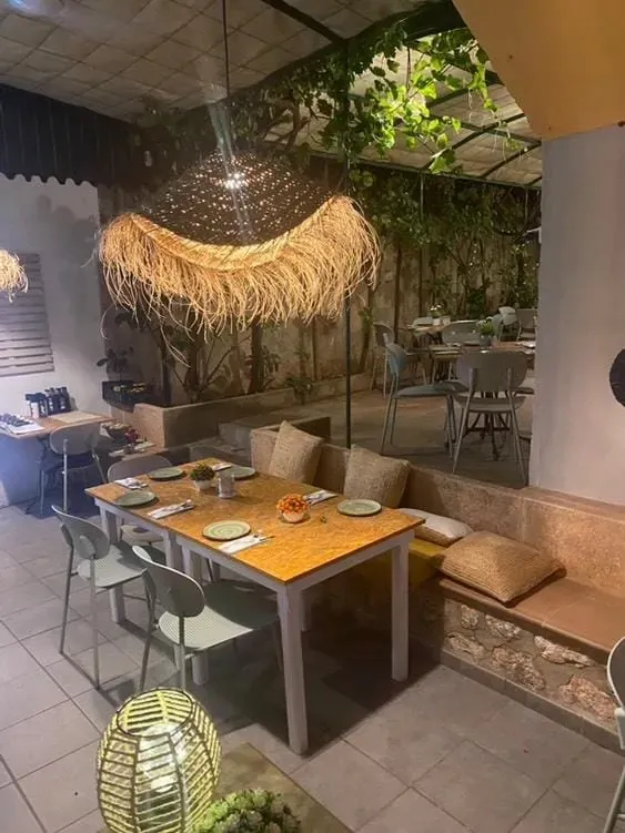 Interior de restaurante con mesas, sillas y lámpara de paja decorativa, rodeado de plantas.