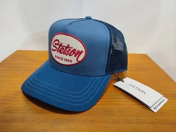 Sombrero camionero Stetson azul con logo rojo y etiqueta blanca sobre una mesa de madera.