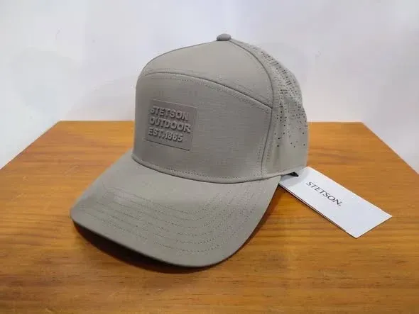 Gorra de béisbol gris con un parche negro en la parte delantera y una etiqueta blanca, sobre una mesa de madera.