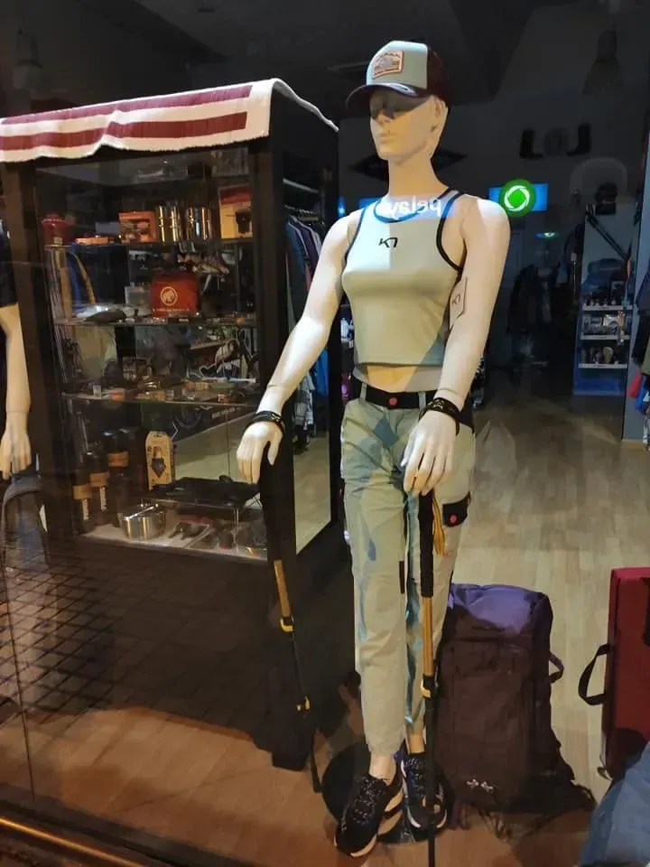 Maniquí con gorra y top corto de pie en el escaparate de una tienda junto a percheros y bolsos.