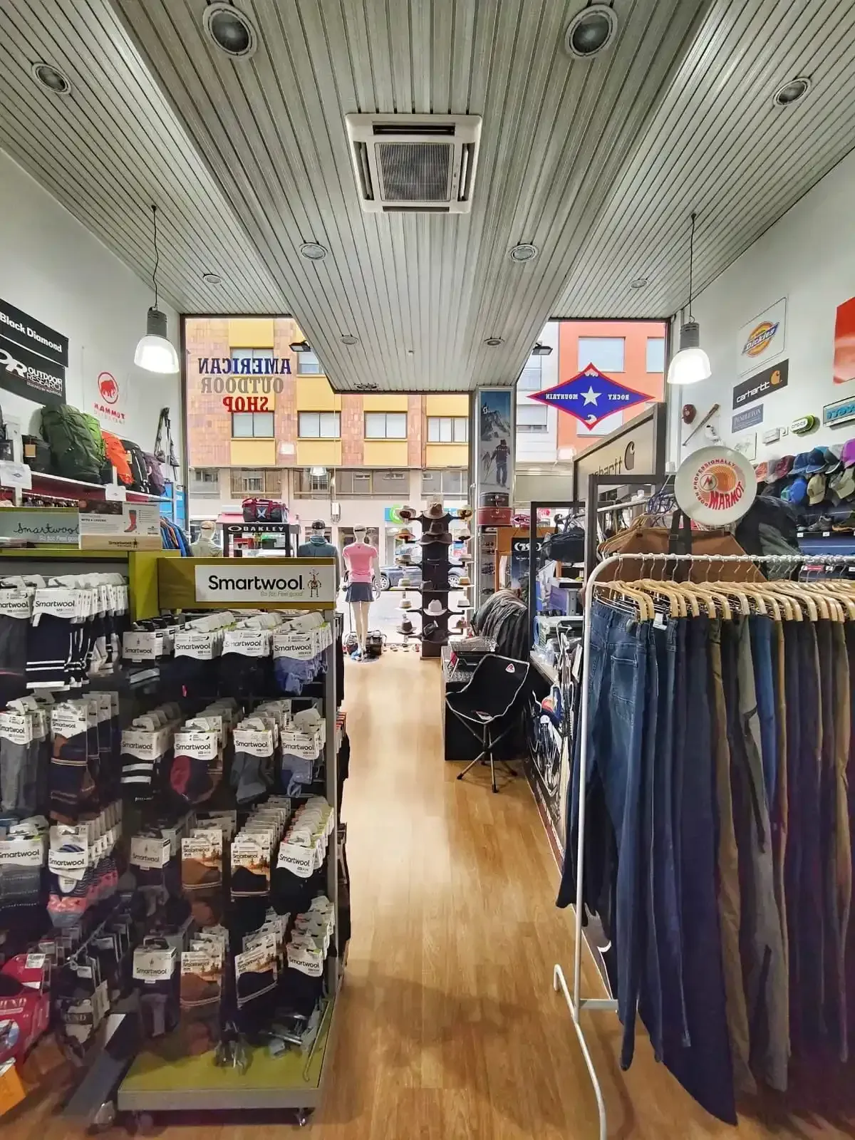 Pasillo de tienda de ropa con percheros de prendas, calcetines y suelo de madera bajo un techo luminoso.