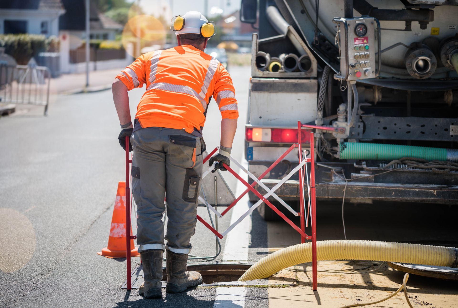Un ouvrier routier en tenue de sécurité orange place une barrière près d'un camion de réparation de voirie et d'un cône de signalisation.