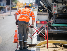 Un ouvrier du bâtiment portant un gilet de sécurité orange dirige la circulation à côté d'engins de chantier et de cônes de signalisation.