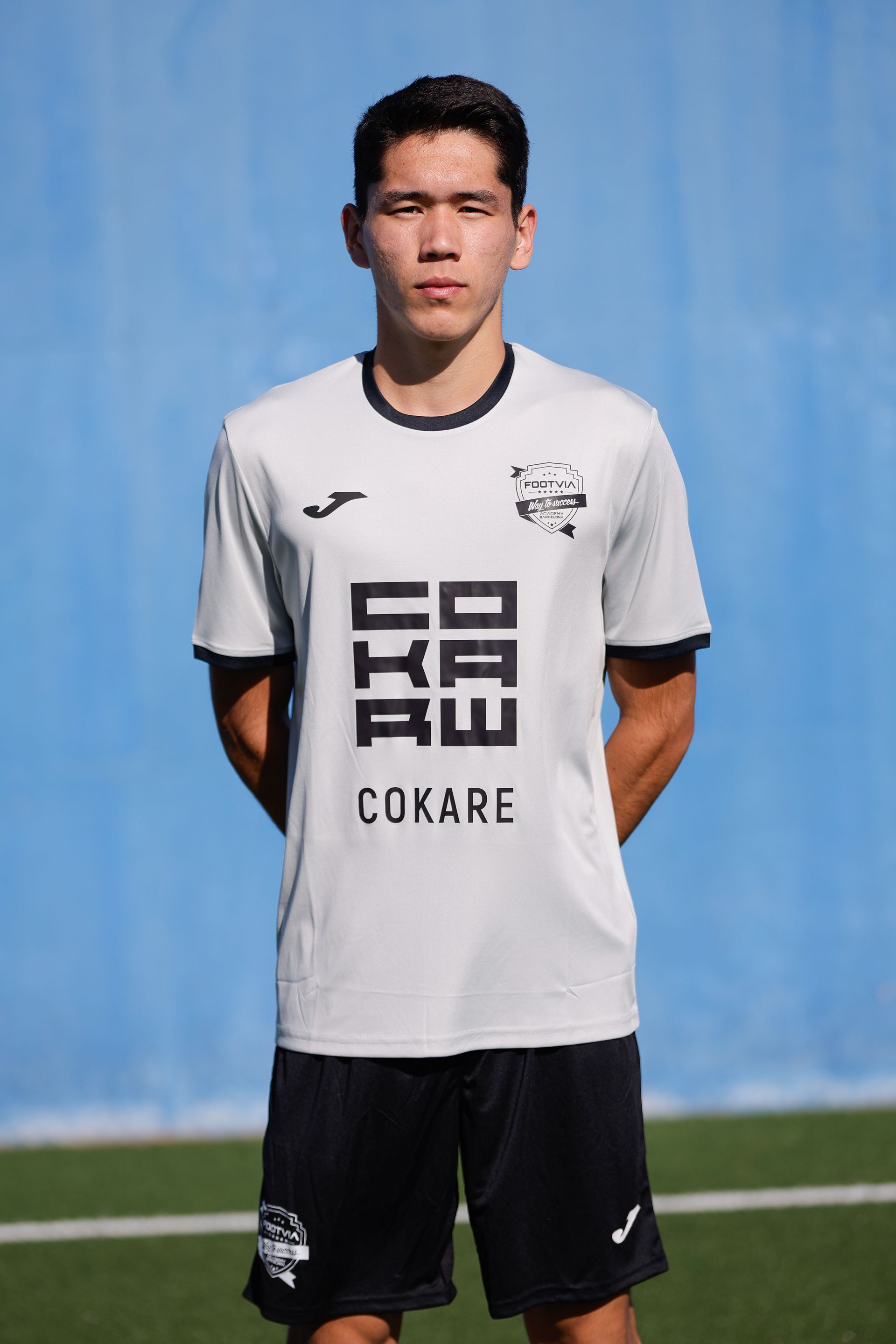 Un jugador de fútbol con una camiseta gris claro con un logotipo oscuro y pantalones cortos negros posa sobre un fondo azul.