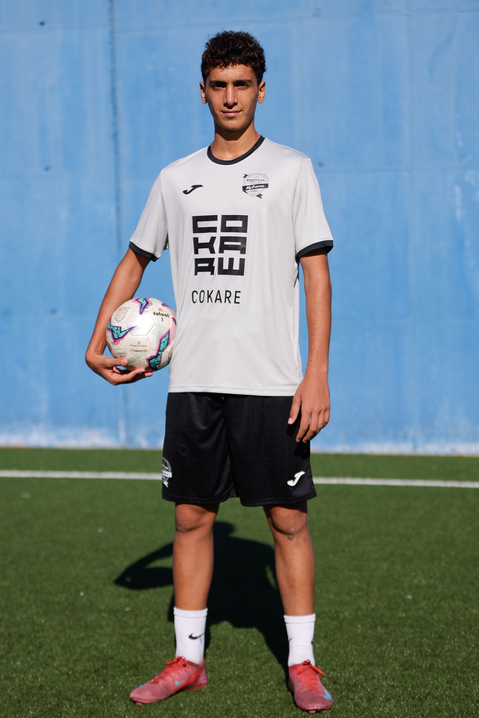 Un jugador de fútbol, ​​vestido con una camiseta blanca y pantalones cortos negros, se encuentra de pie sobre un campo de césped artificial, sosteniendo un balón de fútbol.