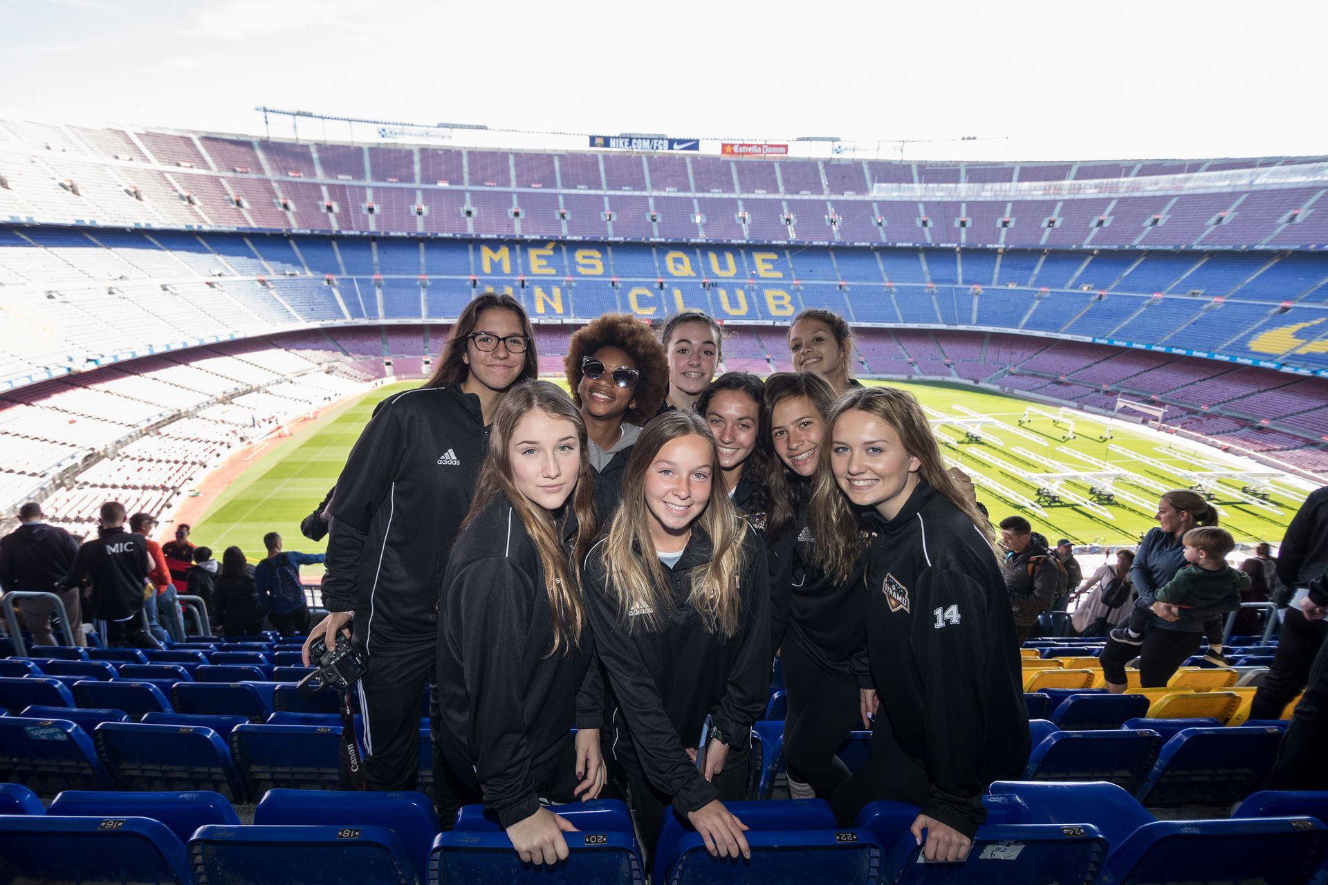 Grupo de mujeres sonriendo frente a un gran estadio, asientos azules y un campo verde visible.
