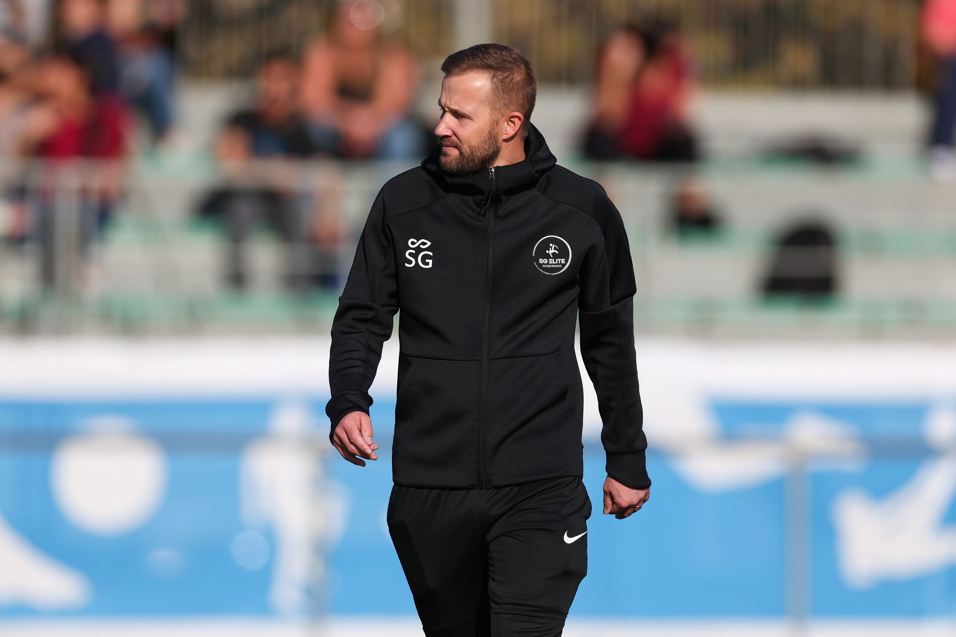 Un entrenador con un chándal negro con capucha camina por un campo deportivo con una multitud sentada al fondo.