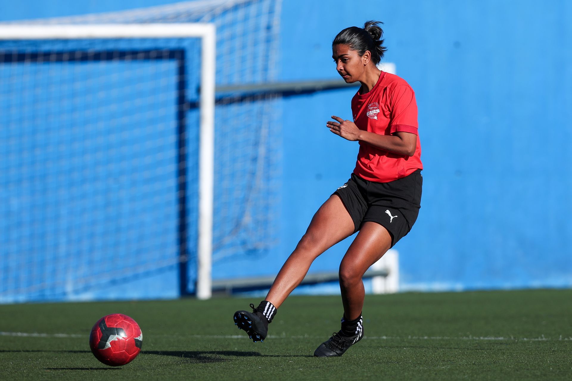Un jugador de fútbol con camiseta roja y pantalones cortos negros patea una pelota roja y blanca en un campo verde.